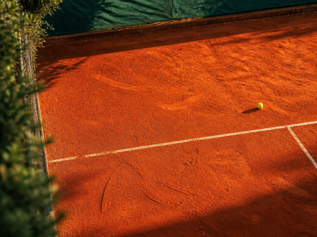 A wide overhead aerial shot of a vivid red European clay tennis court bathed…