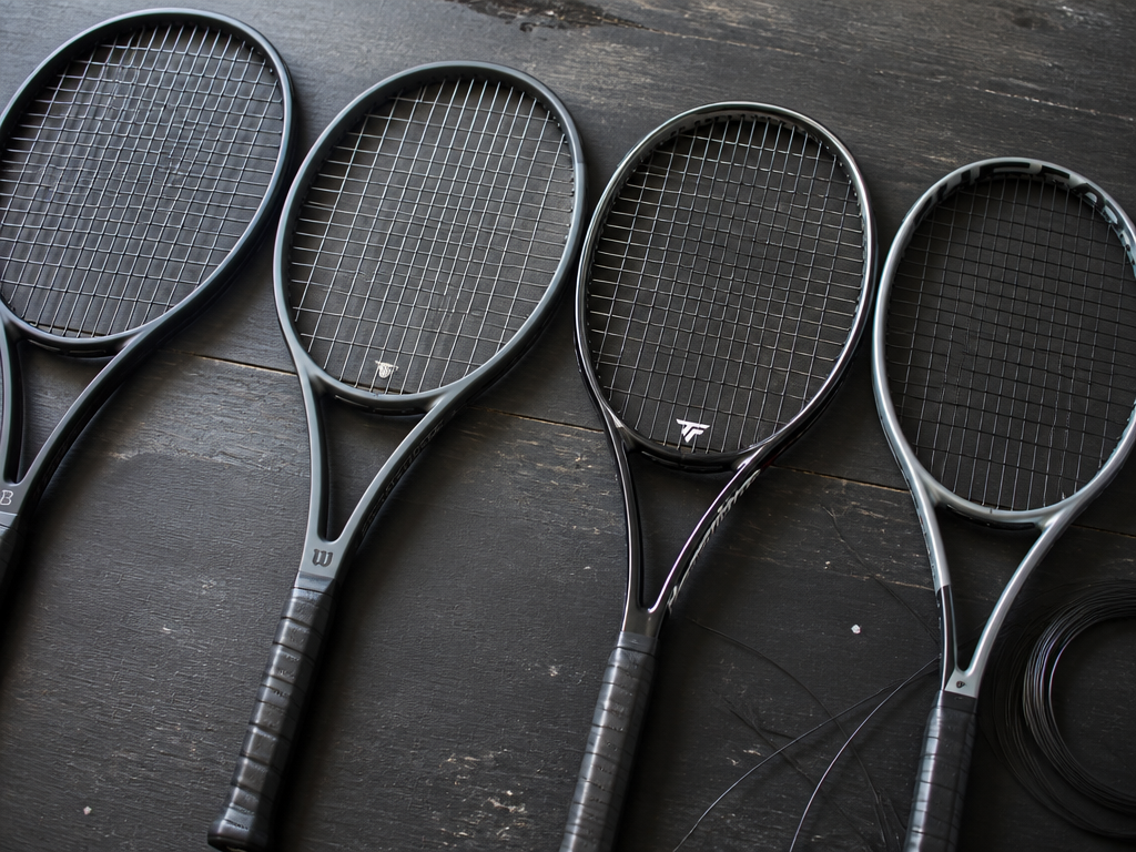 A close-up overhead photograph of four modern tennis racquets laid side by side on…