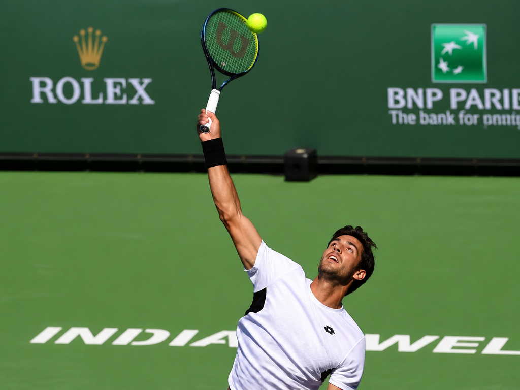 A dynamic action photograph of a right-handed tennis player mid-serve on a hard court…